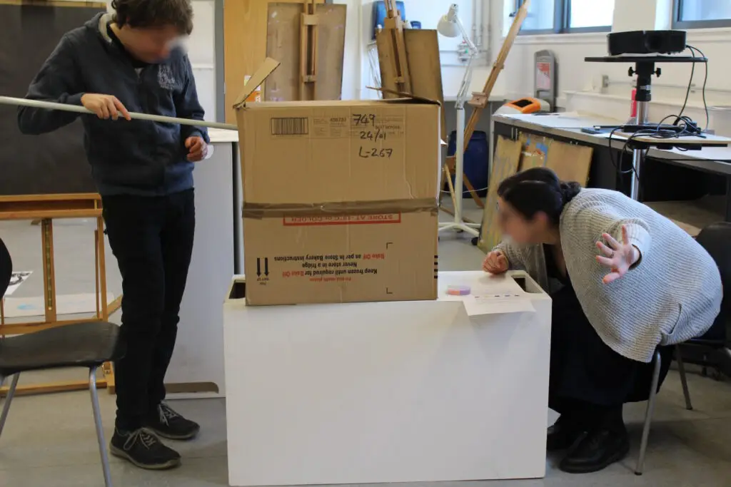 Two playtest participants stand either side of a large cardboard box on a table. One participant is holding a fishing pole, which they have stuck into the side of the box. The other participant crouches down to peer into a hole on their side of the box, and sticks their hand out to the left, to direct the first participant on where to angle the fishing pole. Both participants faces are blurred.