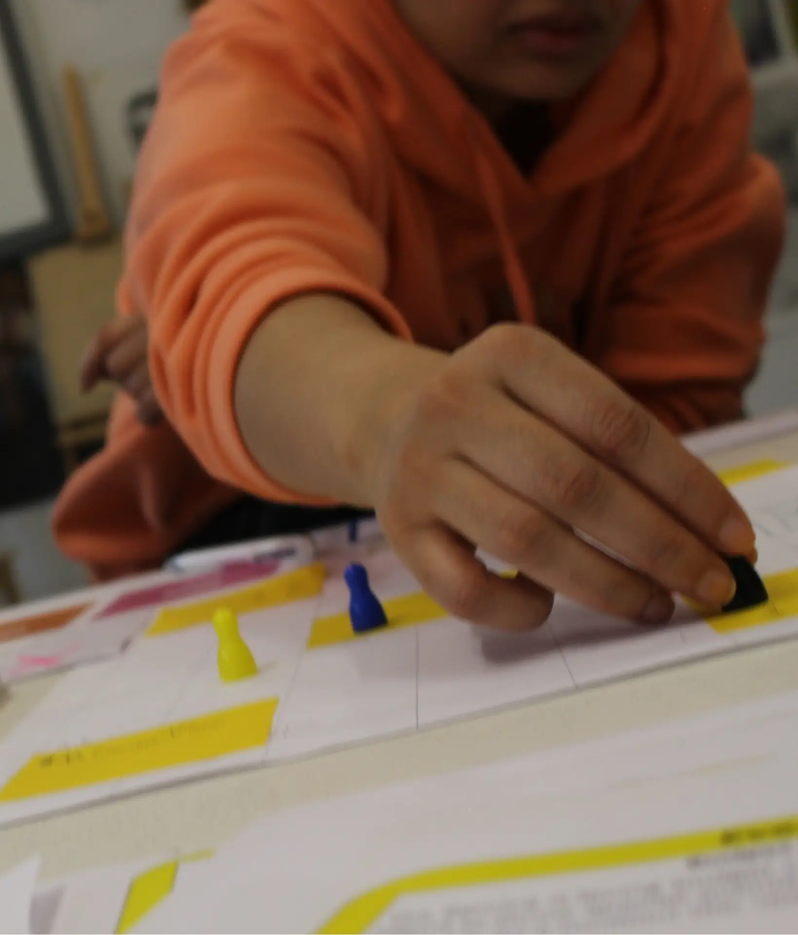 Close-up on a playtest participant's hand reaching out to pick up a meeple. The meeple is on a yellow and white prototype paper game board. Also on the game board are two other meeples. The participant's face has been cropped out of the photo, but they are wearing an orange hoodie.