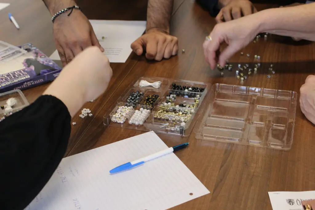 A close-up of five hands gathered around a table, picking lettered beads out of a plastic pack of beads in the centre of the table and sorting them into groups. One group is a pile of small white circular beads showing the letters "I" and "T". The other is a group of gold and silver cubed beads; from the angle of the photograph it is not clear what letters are written on these beads.