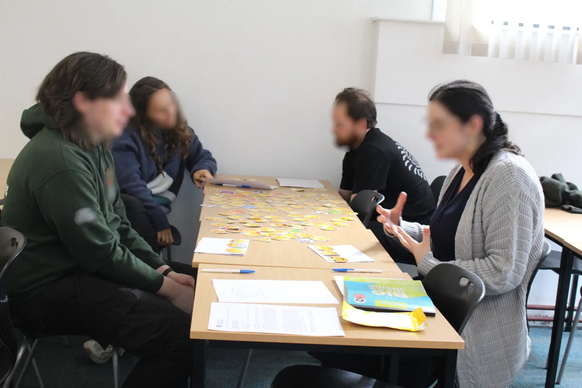 Four playtest participants sit around a long table, speaking animatedly. Spread out on the table is a large collection of stickers, each of a different "emoji". Also on the table are notepads and sheets of paper.