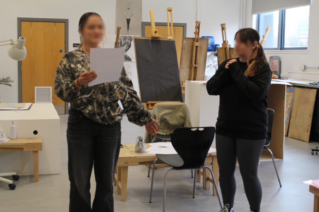 Two playtesters stand in a room. One holds a piece of paper and reads off of it, holding out her free hand in a dramatic gesture. The other watches her, hands clasped together in front of her mouth as she beatboxes. Both participants' faces have been blurred.