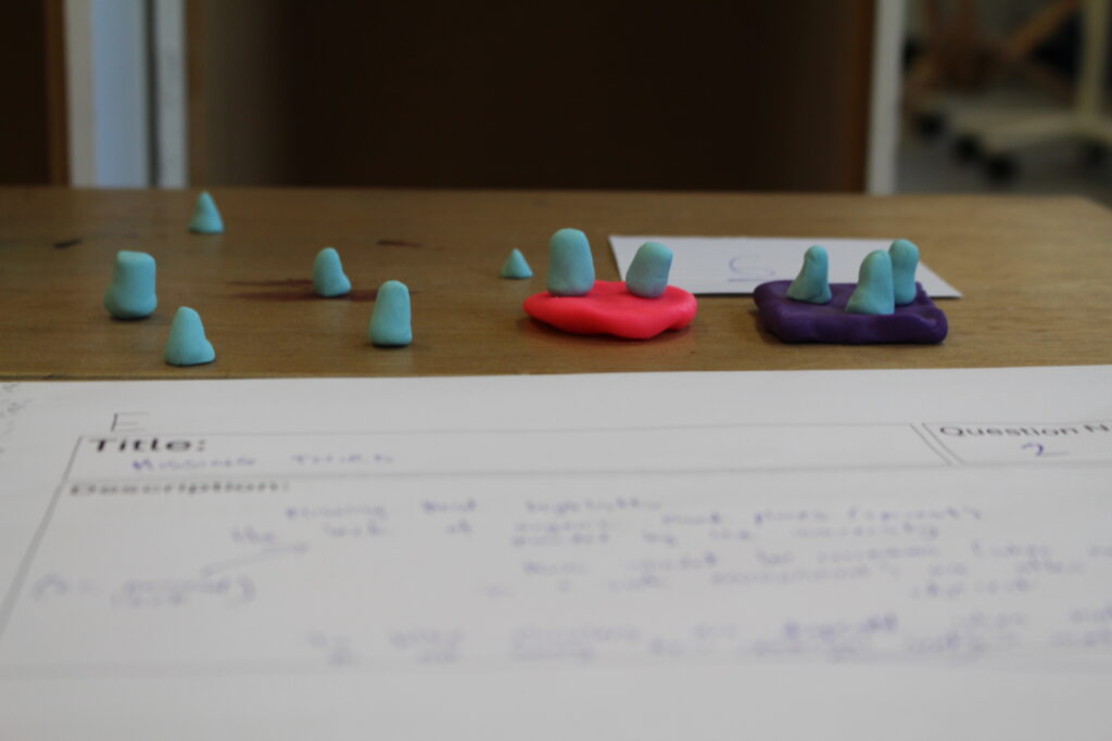 A side-on view of a playtest participant's plasticine sculpture. Three small blue plasticine blocks sit on a purple plasticine square; next to them, two blue blocks sit on a similar pink square. Six more blue blocks are spread out across the table at random. In the foreground is a sheet of paper, on which has been written an explanation of the sculpture, although the writing is not legible from this angle.