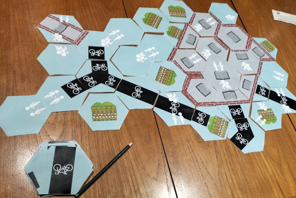 A photograph of a collection of paper hexagon tiles arranged on a table. The tiles have a mixture of symbols on them: two stick figures talking; a cycle path; a garden; a book closed in by walls; and one stick figure helping another stand up. Next to the tableau is a pile of more tiles, and a pencil.